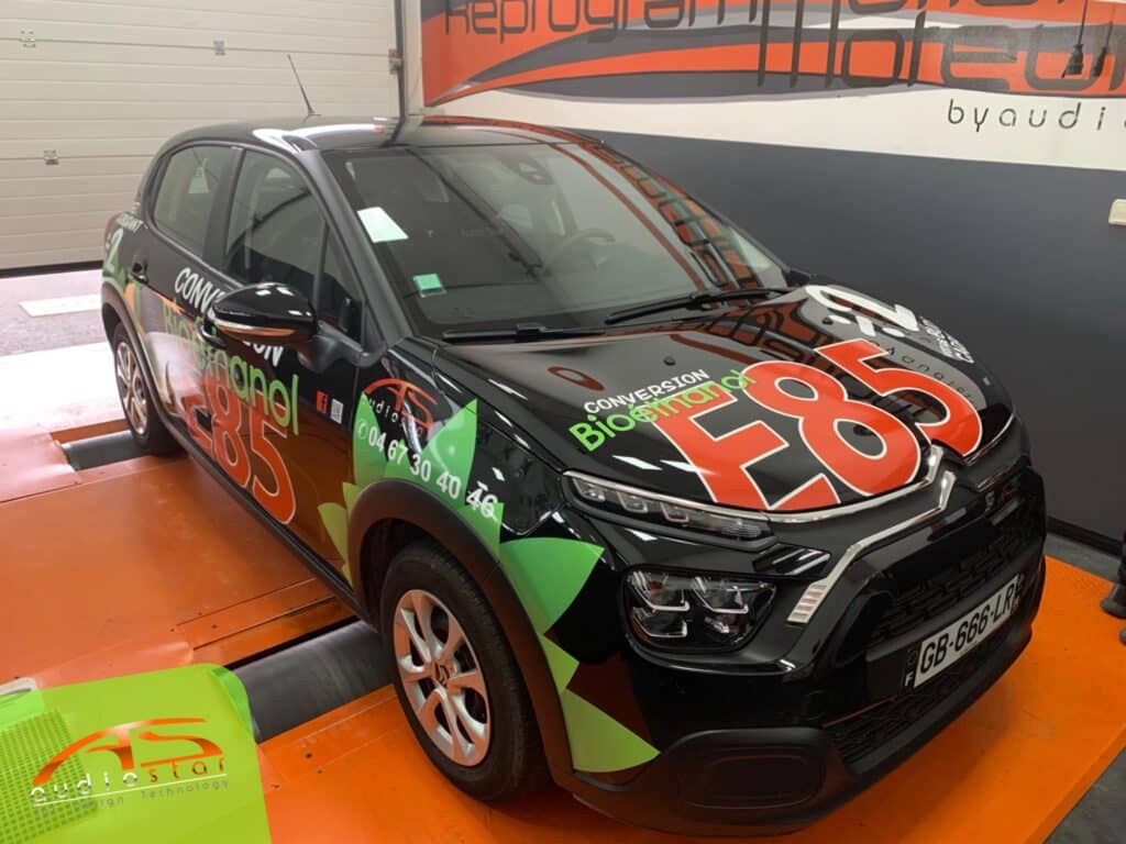 Voiture noire marquée Conversion Bioethanol E85 et Reprogrammation Audiostar. Numéro 04 67 30 40 46 visible. Sur stand d'atelier.