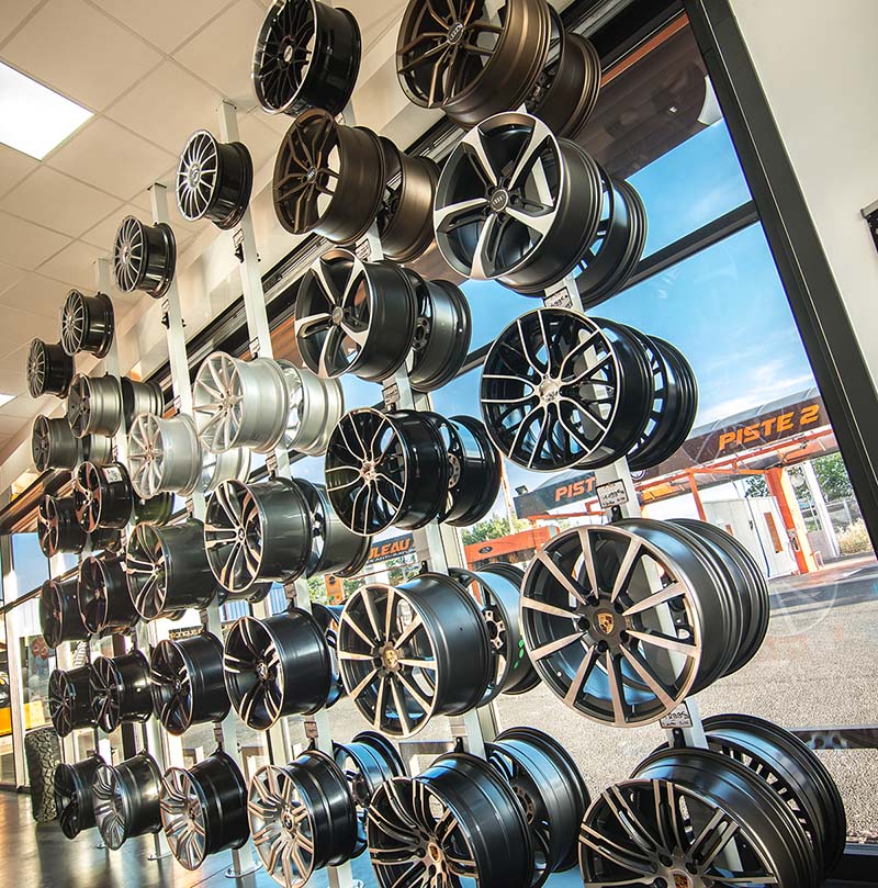 Présentation de nombreuses jantes de voiture sur un mur de magasin. Vue extérieure sur une station de lavage auto avec "PISTE 2".