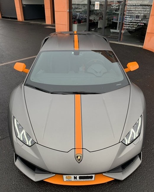 Lamborghini Huracan gris mat, bande orange vif. Lamborghini Huracan gris mat avec bande orange, rétros orange. Plaque AND N. Devant un garage auto offrant diagnostic et reprogrammation.